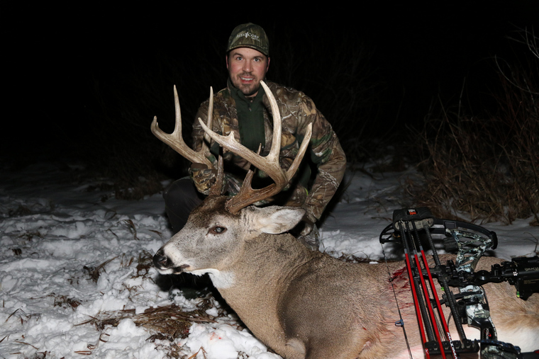 Hunter proudly displaying an 8-point deer, in the snow showcasing the successful harvest from a bow hunt.