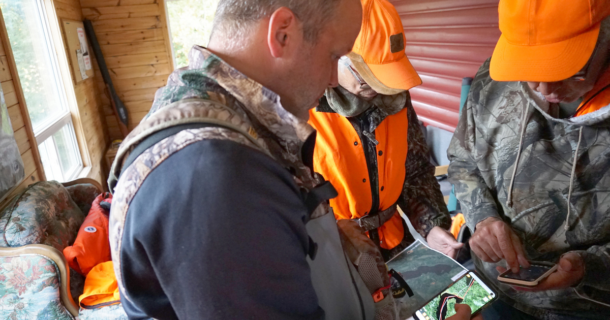 Three hunters in camp wearing camo and hunter orange, gathered around a map and a digital device, planning their hunt.