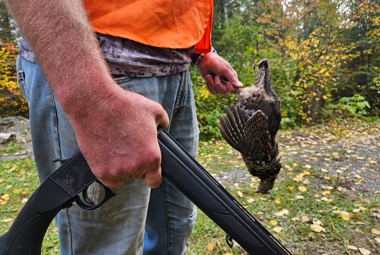 A hunter holding a grouse in one hand and a break-action 28-gauge shotgun in the other, showcasing a successful hunt.