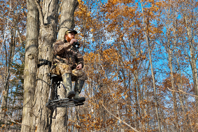 A camouflaged hunter sitting in a climbing tree stand, holding binoculars and scanning the surroundings