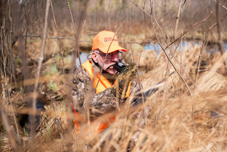 A hunter wearing hunter orange and camo sits in the heart of a swamp, calling for deer with focused attention.