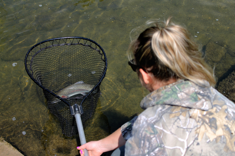 Over-the-shoulder view of an angler holding a net with a freshly caught rainbow trout on a creek