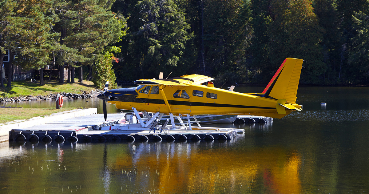 A yellow floatplane tied to a dock at the edge of a calm lake, bathed in sunlight on a clear summer day.