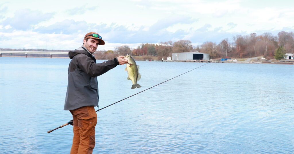 Shore angler proudly shows off a freshly caught largemouth bass with a scenic lake behind.
