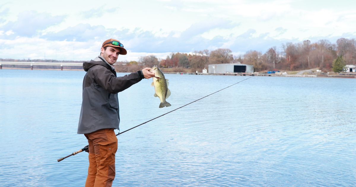 Shore angler proudly shows off a freshly caught largemouth bass with a scenic lake behind.
